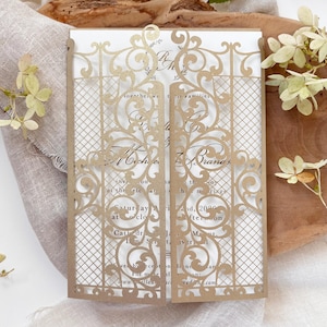 May include: A gold-colored wedding invitation with a laser-cut gate design. The invitation features intricate floral and scroll patterns. The text on the invitation includes names, date, time, and location details. The invitation is displayed on a wooden surface.