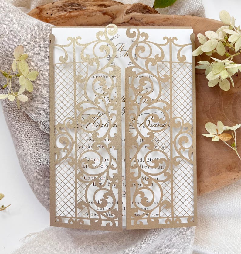 May include: A gold-colored wedding invitation with a laser-cut gate design. The invitation features intricate floral and scroll patterns. The text on the invitation includes names, date, time, and location details. The invitation is displayed on a wooden surface.