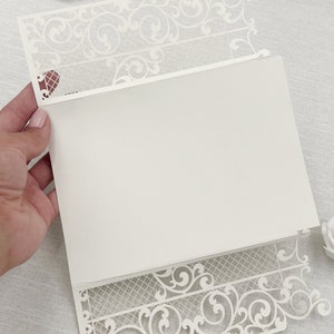 May include: Ivory laser-cut invitation with intricate floral and scrollwork design. The invitation features a decorative border and a blank interior for personalized text. A heart-shaped detail is visible on the upper left corner. Perfect for weddings or special events.