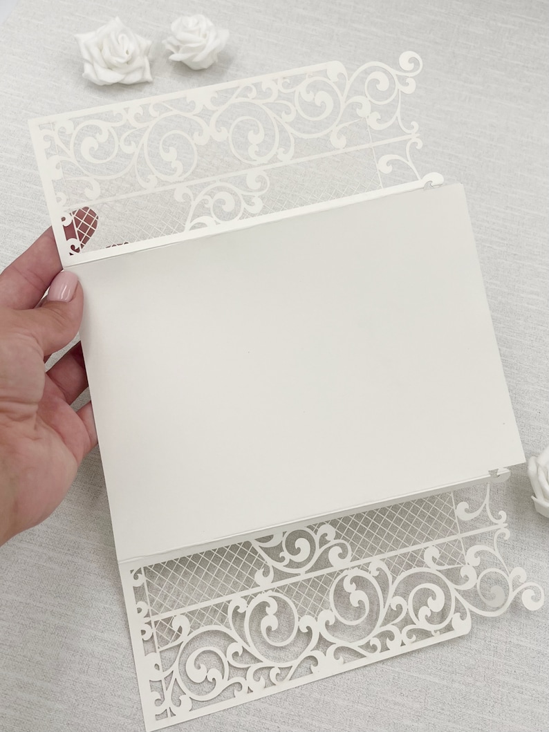 May include: Ivory laser-cut invitation with intricate floral and scrollwork design. The invitation features a decorative border and a blank interior for personalized text. A heart-shaped detail is visible on the upper left corner. Perfect for weddings or special events.