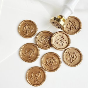 Może przedstawiać: Złote pieczęcie woskowe z monogramami, rozrzucone na białej powierzchni. Złoty stempel woskowy z białym uchwytem jest umieszczony nad pieczęciami. Pieczęcie zawierają skomplikowane kombinacje liter, idealne do papeterii.