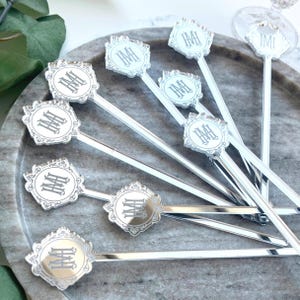 May include: Silver cocktail stirrers with a decorative, ornate design. Each stirrer features a circular emblem with the initials "IM" in a stylized font. The stirrers are arranged on a marble-patterned tray, suggesting a bar or entertaining setting.