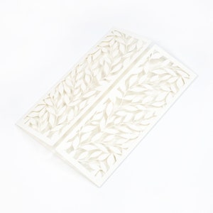 May include: White wedding invitation with a delicate, intricate design of leaves cut out of the card stock.