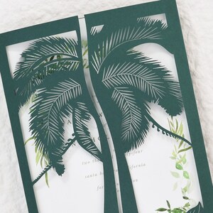 May include: Dark green laser-cut invitation with a palm tree design. The invitation has a white interior with watercolor greenery and text. The invitation is open, revealing the interior details.