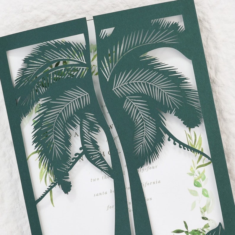 May include: Dark green laser-cut invitation with a palm tree design. The invitation has a white interior with watercolor greenery and text. The invitation is open, revealing the interior details.