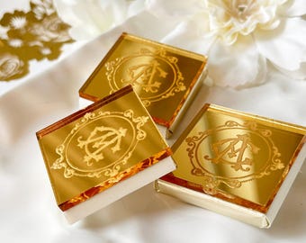 May include: Three square gold mirror acrylic plaques featuring an engraved ornate frame surrounding the intertwined letters AM. Each reflective piece is mounted on a thick white base, creating a layered effect with a polished metallic finish.