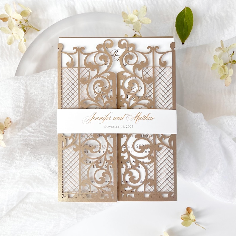 May include: A laser-cut wedding invitation in a gate design. The invitation is a light brown color with intricate scrollwork and a lattice pattern. The names "Jennifer and Matthew" and the date "November 1, 2025" are printed on a white band.
