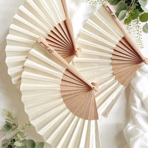 Puede incluir: Varios abanicos de papel color crema con mangos de madera están dispuestos sobre una superficie blanca. Los abanicos están desplegados, mostrando su diseño plisado. Los mangos de madera tienen texto. Hay vegetación alrededor de los abanicos.