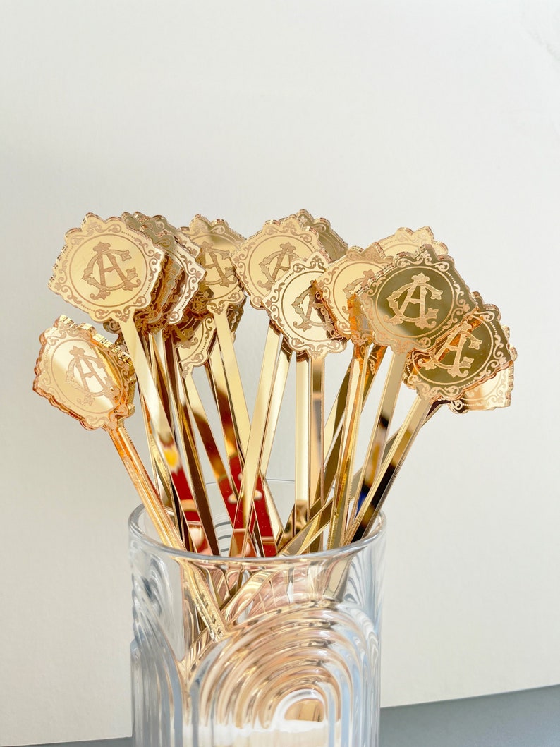 May include: A bunch of gold-colored metal cocktail stirrers with a decorative, ornate, monogrammed design. The stirrers are in a clear glass vase.