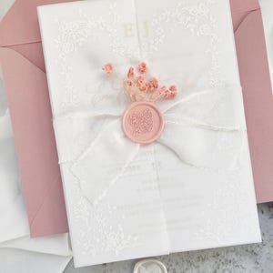Puede incluir: Una invitación de boda blanca con detalles de encaje floral, atada con una cinta blanca y sellada con un sello de cera rosa. La invitación está en un sobre rosa, con dos sellos de cera adicionales en la parte inferior.