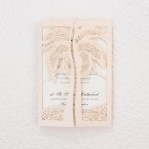 May include: A light peach-colored wedding invitation with a laser-cut palm tree design. The invitation is open, revealing printed text with event details. The invitation is on a white textured surface.