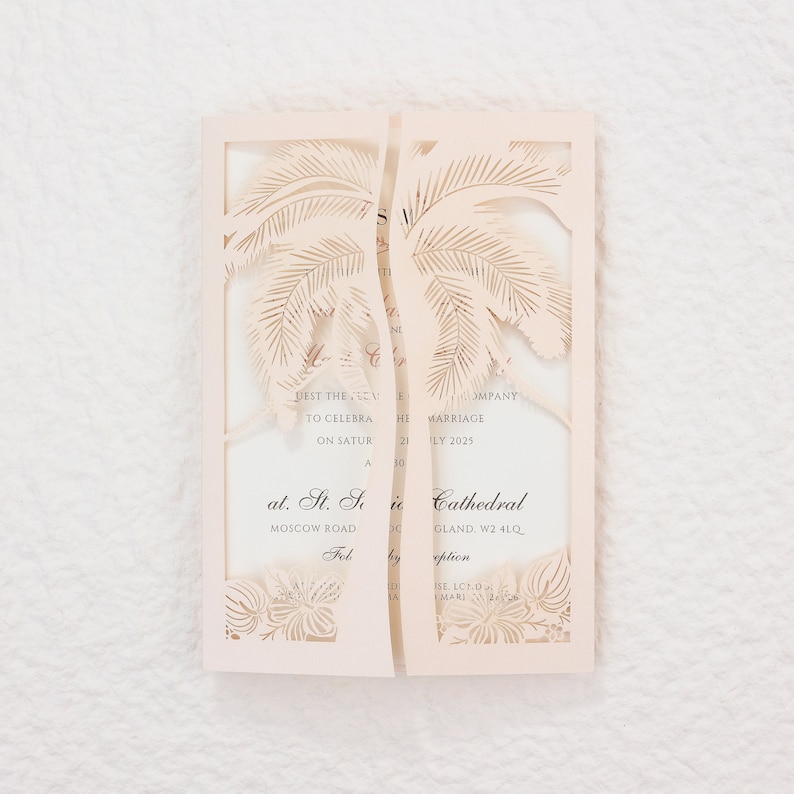 May include: A light peach-colored wedding invitation with a laser-cut palm tree design. The invitation is open, revealing printed text with event details. The invitation is on a white textured surface.