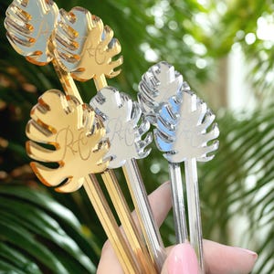 May include: A collection of gold and silver-colored leaf-shaped drink stirrers. Each stirrer features a detailed leaf design and a monogram. The stirrers are held against a backdrop of green foliage.
