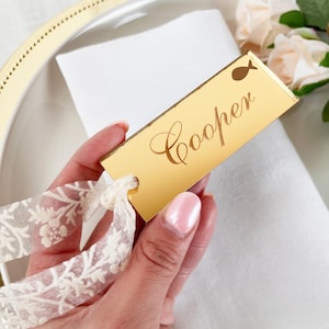 May include: A gold rectangular place card with the name "Cooper" in cursive script. A small fish icon is above the name. The card is attached to a white and cream floral ribbon. The card is placed on a white napkin.
