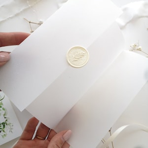Puede incluir: Sobre blanco con un sello de cera blanco con un dise&ntilde;o de hoja. El sobre est&aacute; doblado por la mitad y sostenido por una mano.