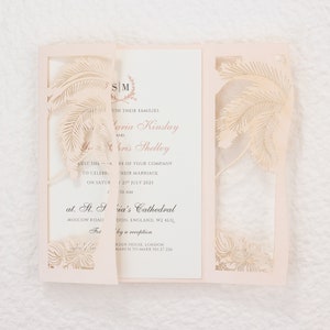May include: A light pink wedding invitation with a laser-cut palm tree design. The invitation is tri-fold, with the text "Maria Kinslay and Chris Shelley" and wedding details printed inside. The invitation includes the date, time, and location of the wedding.