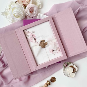 Op de afbeelding: Een met oudroze stof beklede uitnodigingsdoos bevat een witte kaart met roze kersenbloesemillustraties, vastgezet met een wit lint en een gouden waszegel. De set bevat een koperen zegelstempel en diverse kleine witte waszegels.