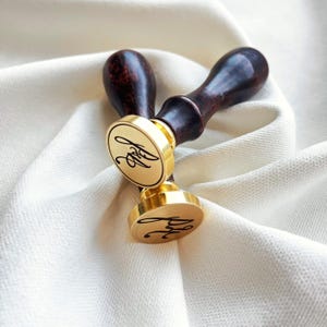 May include: Two gold wax seal stamps with dark brown wooden handles. The stamps feature a circular design with a cursive monogram. They are placed on a white, textured fabric.