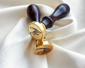 May include: Two gold wax seal stamps with dark brown wooden handles. The stamps feature a circular design with a cursive monogram. They are placed on a white, textured fabric.