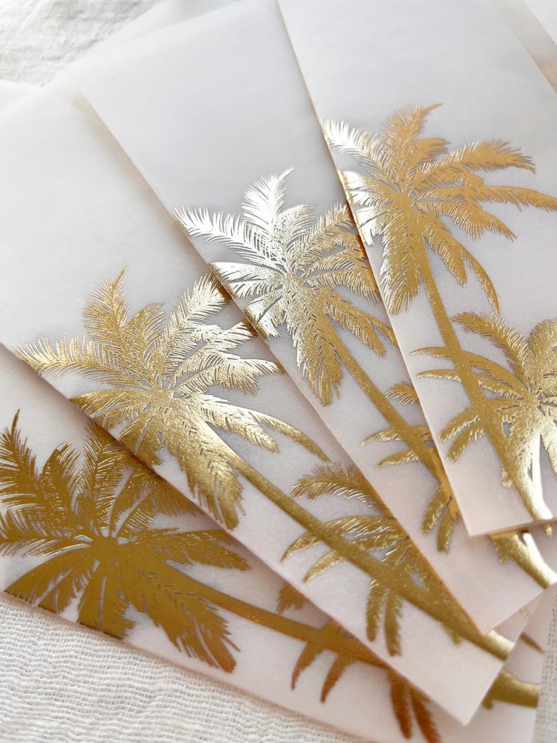 Peut inclure: Une pile de serviettes en papier blanches avec un motif de palmier dor&eacute;. Les palmiers sont d&eacute;taill&eacute;s avec du papier d'aluminium dor&eacute;, cr&eacute;ant un effet scintillant. Les serviettes sont dispos&eacute;es en &eacute;ventail, mettant en valeur le design tropical.