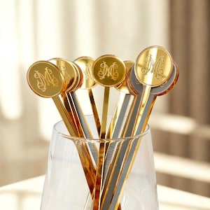 May include: A collection of gold-colored cocktail stirrers with an ornate letter "M" design on the circular tops. The stirrers are displayed in a clear glass, with some featuring a gold and gray color scheme. The background is softly lit.