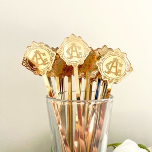 May include: A set of gold-colored metal cocktail stirrers with a decorative, ornate design and a monogrammed initial "A" on each. The stirrers are in a clear glass vase.