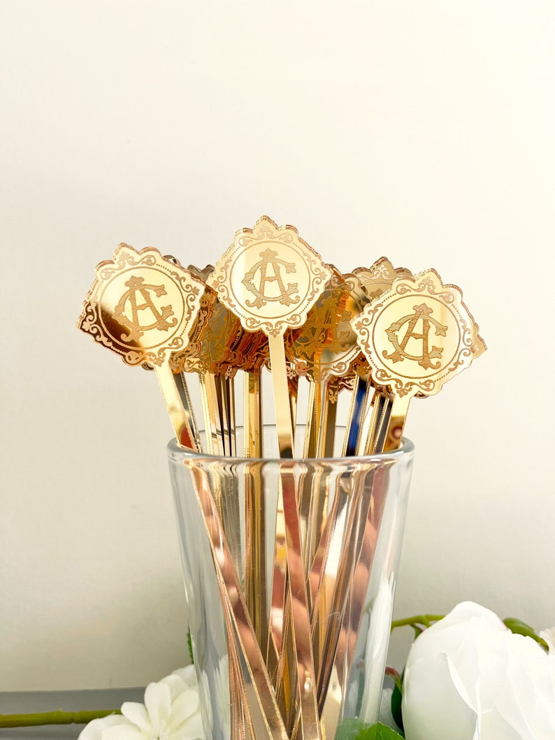 May include: A set of gold-colored metal cocktail stirrers with a decorative, ornate design and a monogrammed initial "A" on each. The stirrers are in a clear glass vase.