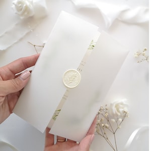 Puede incluir: Invitaci&oacute;n de boda blanca con un sello de cera y detalles florales verdes. La invitaci&oacute;n est&aacute; doblada por la mitad y tiene el texto "we'll be wed" y "11.11" impreso en ella.