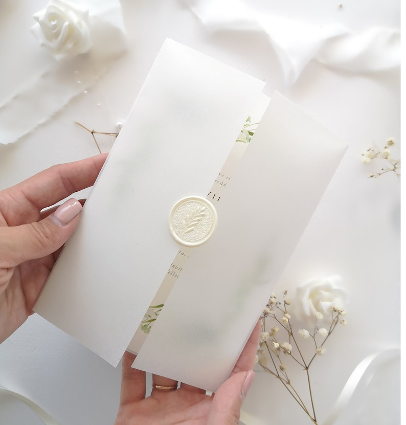 Puede incluir: Invitaci&oacute;n de boda blanca con un sello de cera y detalles florales verdes. La invitaci&oacute;n est&aacute; doblada por la mitad y tiene el texto "we'll be wed" y "11.11" impreso en ella.