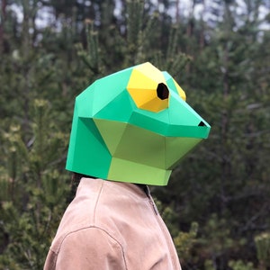 Op de afbeelding: Een groen en geel papieren masker van een kikker. Het masker is gemaakt van gevouwen papier en heeft een driedimensionaal effect.