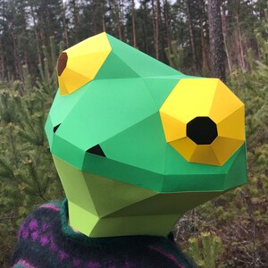 Op de afbeelding: Een groen en geel papieren kikkermasker met zwarte ooggaten. Het masker is gemaakt van gevouwen papier en heeft een 3D-effect.