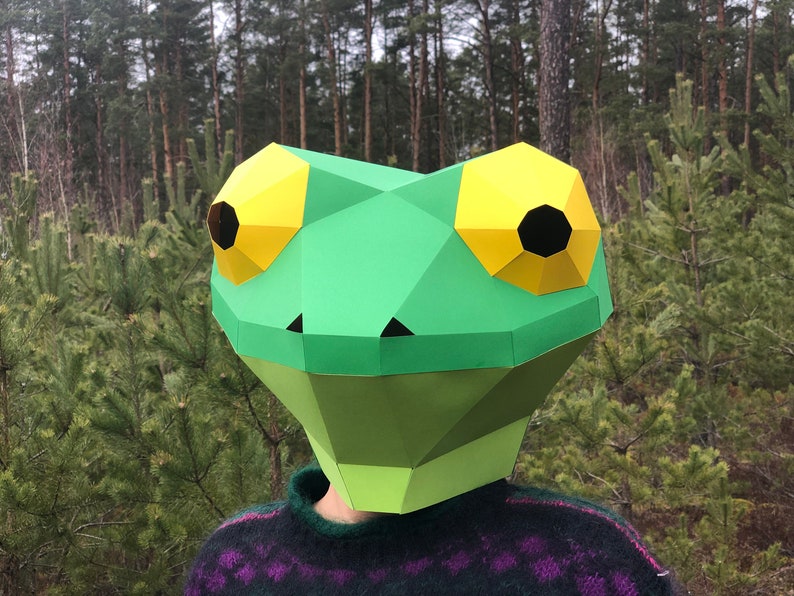 Op de afbeelding: Een groen papieren kikkermasker met gele ogen en zwarte pupillen. Het masker wordt gedragen door een persoon in een bosrijke omgeving.