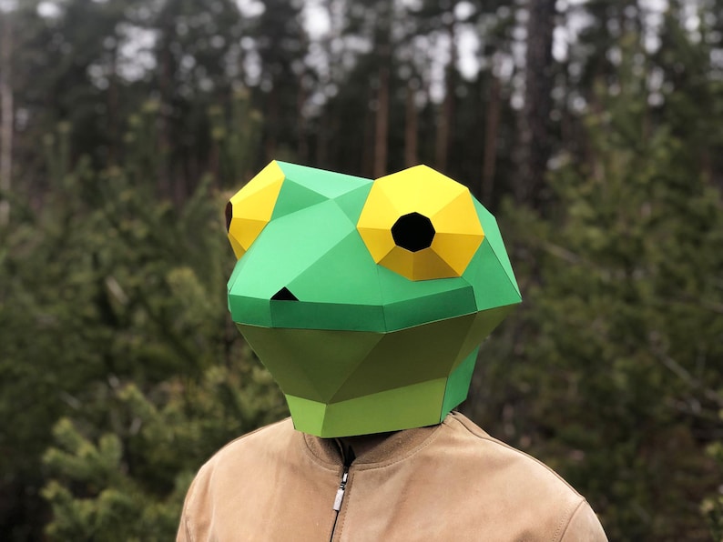 Op de afbeelding: Een groen en geel papier-mach&eacute; kikkermasker. Het masker is gemaakt van gevouwen papier en heeft een driedimensionaal ontwerp.