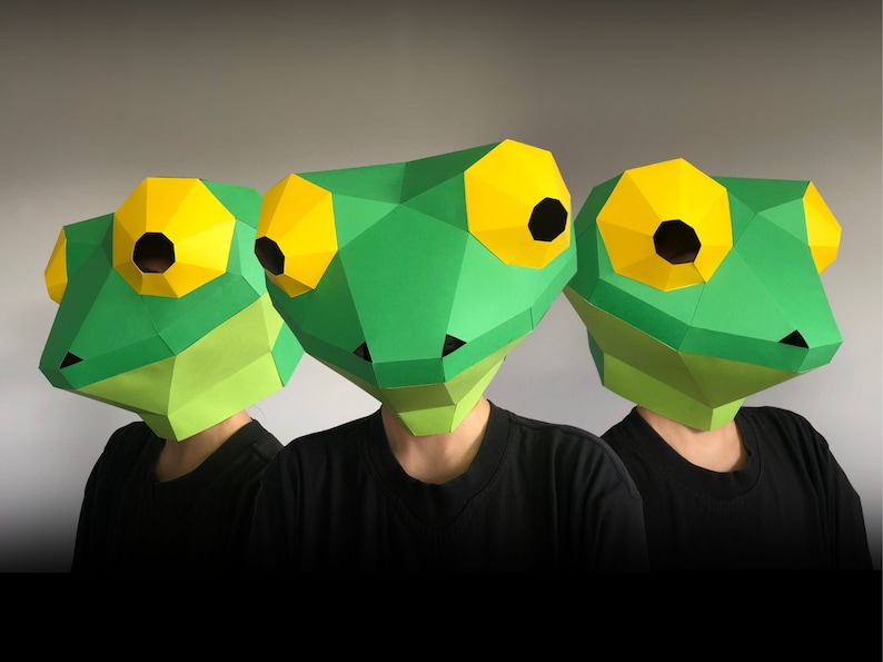 Op de afbeelding: Drie groene papieren maskers van een kikker met gele ogen en zwarte pupillen. De maskers worden gedragen door mensen met zwarte shirts.