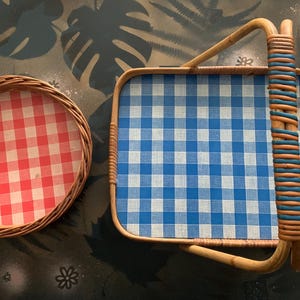 May include: Two wicker trays with gingham patterns. One is round with a red and white pattern, and the other is rectangular with a blue and white pattern. The rectangular tray has a woven handle.