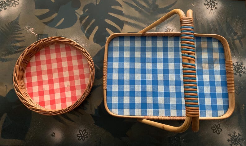 May include: Two wicker trays with gingham patterns. One is round with a red and white pattern, and the other is rectangular with a blue and white pattern. The rectangular tray has a woven handle.