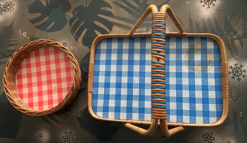 May include: Two vintage picnic baskets. One is round with a red and white checkered pattern, and the other is rectangular with a blue and white checkered pattern. Both have woven wicker frames.