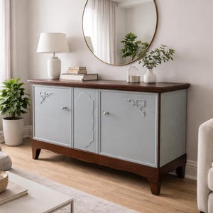 Könnte beinhalten: Hellblau-braune Holz-Anrichte mit drei Türen und silberfarbenen Griffen. Die Oberseite ist dunkelbraun, die Türen hellblau mit dekorativen Akzenten. Ein runder Spiegel hängt über der Anrichte.
