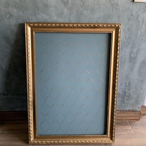 Könnte beinhalten: Ein rechteckiger goldfarbener Bilderrahmen mit aufwendigen Details. Der Rahmen umgibt eine blaue Oberfläche mit einem goldenen geometrischen Muster. Der Rahmen liegt auf einem braunen Holzboden vor einer grauen Wand.