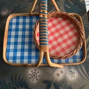 May include: A vintage picnic basket with a woven handle and frame. The basket features a blue and white checkered pattern on the base and a red and white checkered pattern on the round tray. The handle is wrapped with blue and orange accents.