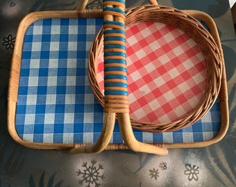 May include: A vintage picnic basket with a woven handle and frame. The basket features a blue and white checkered pattern on the base and a red and white checkered pattern on the round tray. The handle is wrapped with blue and orange accents.