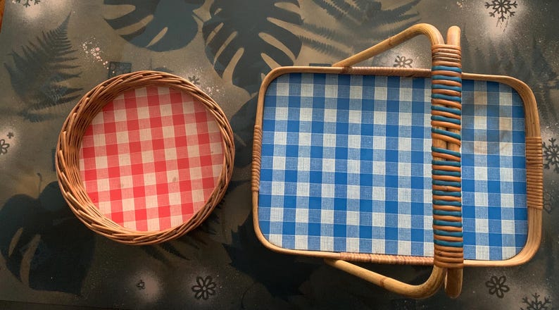 May include: Two wicker serving trays. One is round with a red and white checkered pattern. The other is rectangular with a blue and white checkered pattern and a handle. The handle is wrapped with blue and brown material.