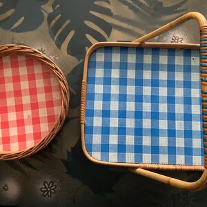 May include: Two vintage wicker trays. One is round with a red and white checkered pattern. The other is rectangular with a blue and white checkered pattern and a woven handle. The trays are made of natural-colored wicker.