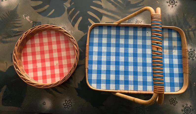 May include: Two vintage wicker trays. One is round with a red and white checkered pattern. The other is rectangular with a blue and white checkered pattern and a woven handle. The trays are made of natural-colored wicker.