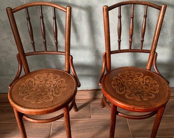May include: Two antique wooden chairs with curved backs and round seats. The seats feature an ornate, light-colored design. The chairs have slender legs and a warm, brown finish, suggesting a vintage aesthetic. The background is a muted green wall.