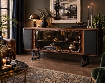 May include: A long wooden sideboard featuring a central glass-fronted cabinet with ornate carved wood detailing, flanked by two solid black side panels. The unit is supported by black metal legs and contains interior shelves holding books and decorative items.