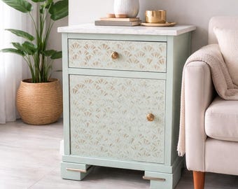 May include: A mint green nightstand with a marble top and gold hardware. The drawer and door feature a fan-shaped pattern. A white vase with flowers, a gold cup, and books sit on top. A large potted plant and a round jute rug are in the foreground.