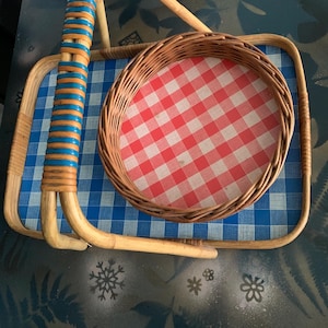 May include: A vintage picnic basket with a blue and white checkered tray and a round basket with a red and white checkered pattern. The handle is made of woven bamboo with blue and orange accents.