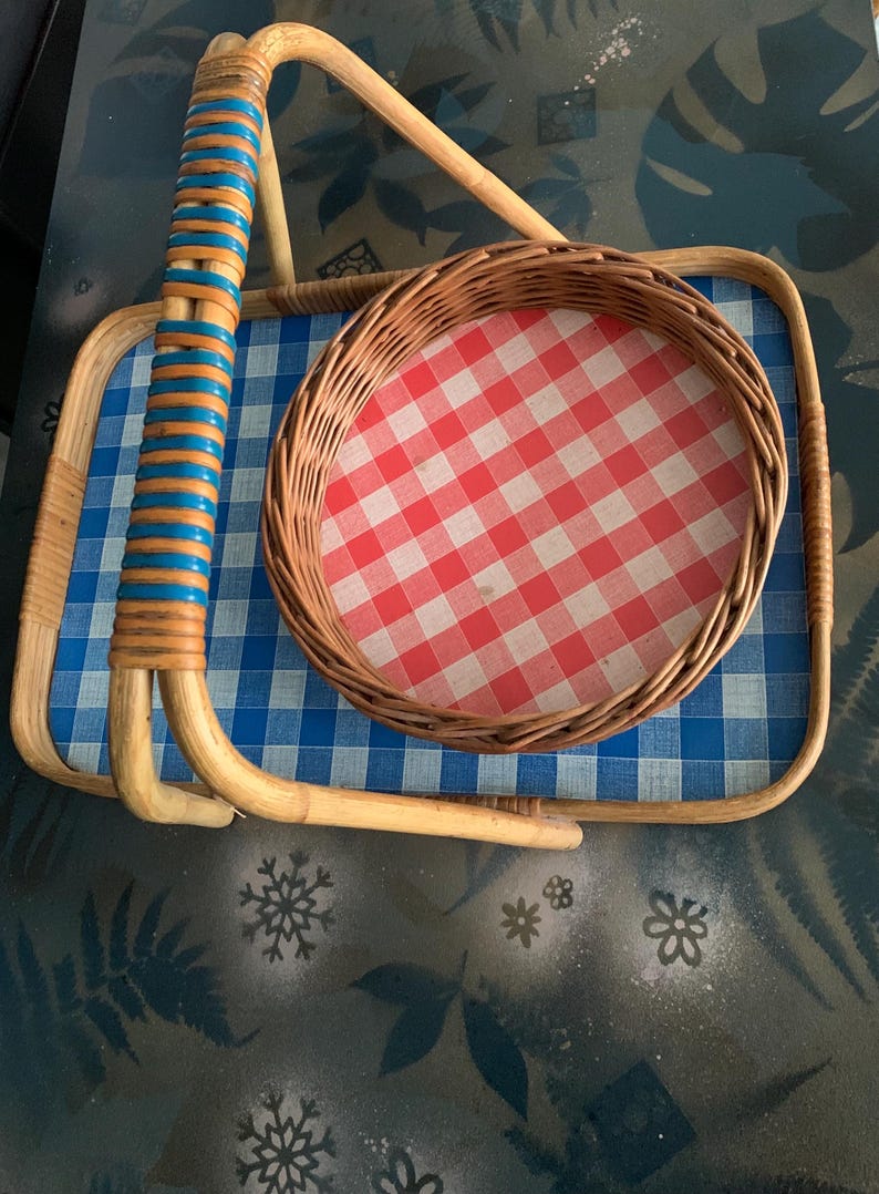 May include: A vintage picnic basket with a blue and white checkered tray and a round basket with a red and white checkered pattern. The handle is made of woven bamboo with blue and orange accents.