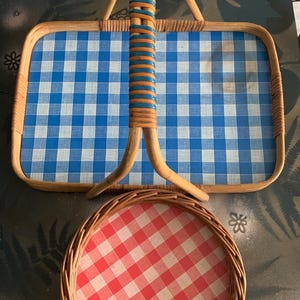 May include: Two picnic baskets. The larger basket is rectangular with a blue and white checkered pattern and a woven handle with blue and brown accents. The smaller basket is round with a red and white checkered pattern.